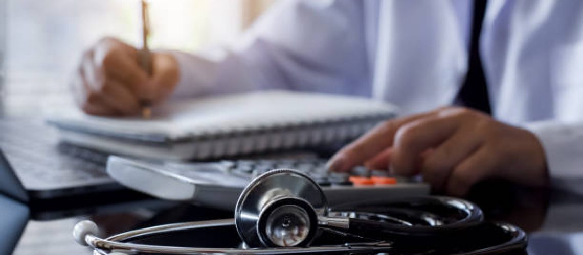 Doctor using calculator and work on laptop computer. Medical cost and fees concept.